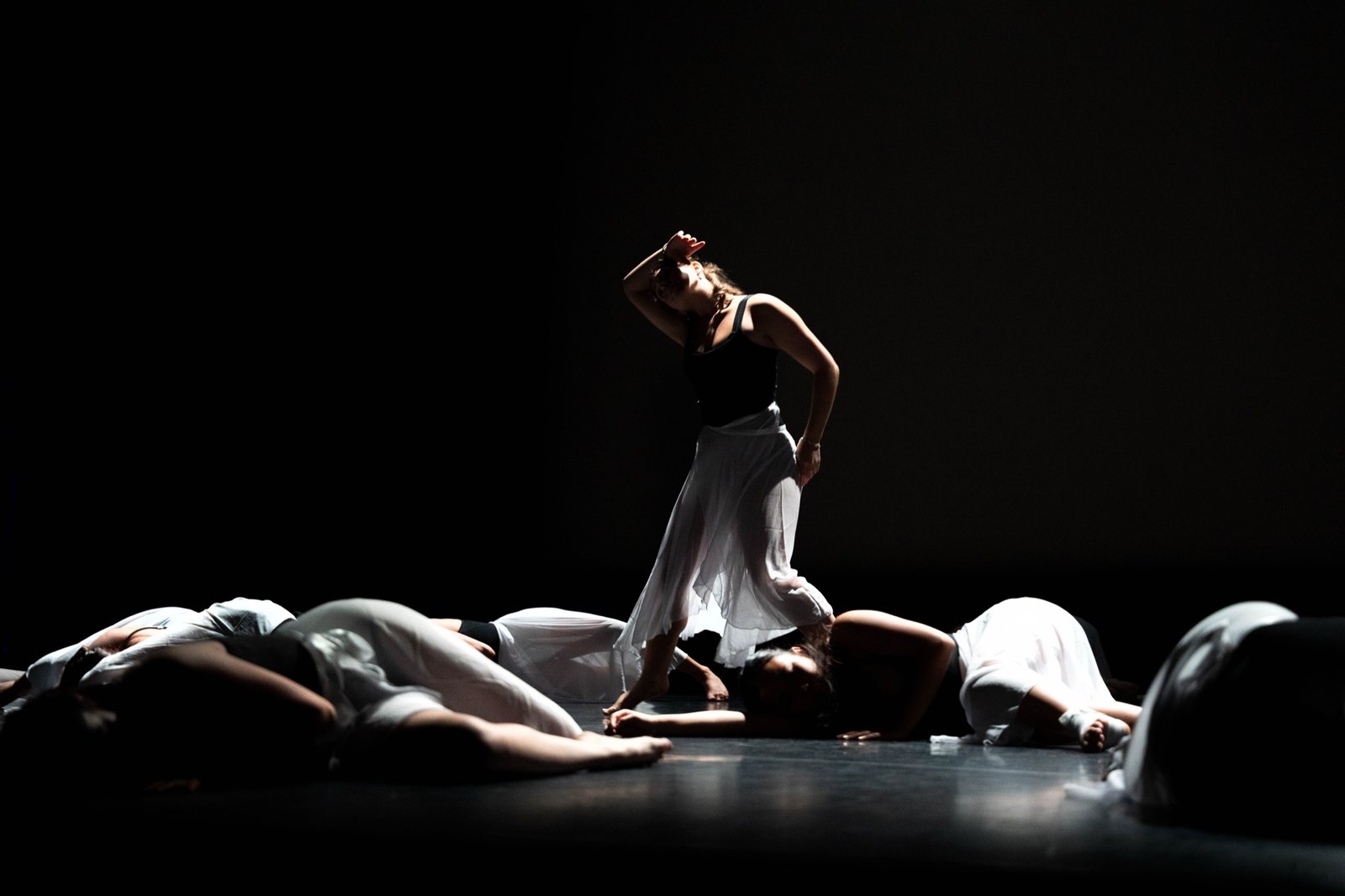 Contemporary dancer lit on a dark stage, other dancers lying on the floor around her
