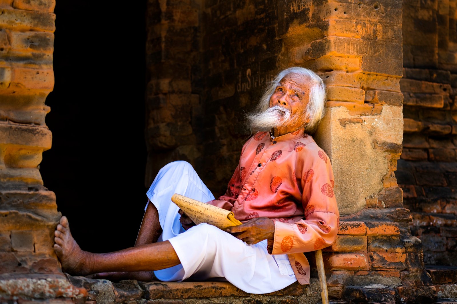 Elderly barefoot man reading, leaning against an ancient brick temple wall